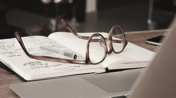 Close up of some glasses and a pen on top of an open notebook in front of a laptop
