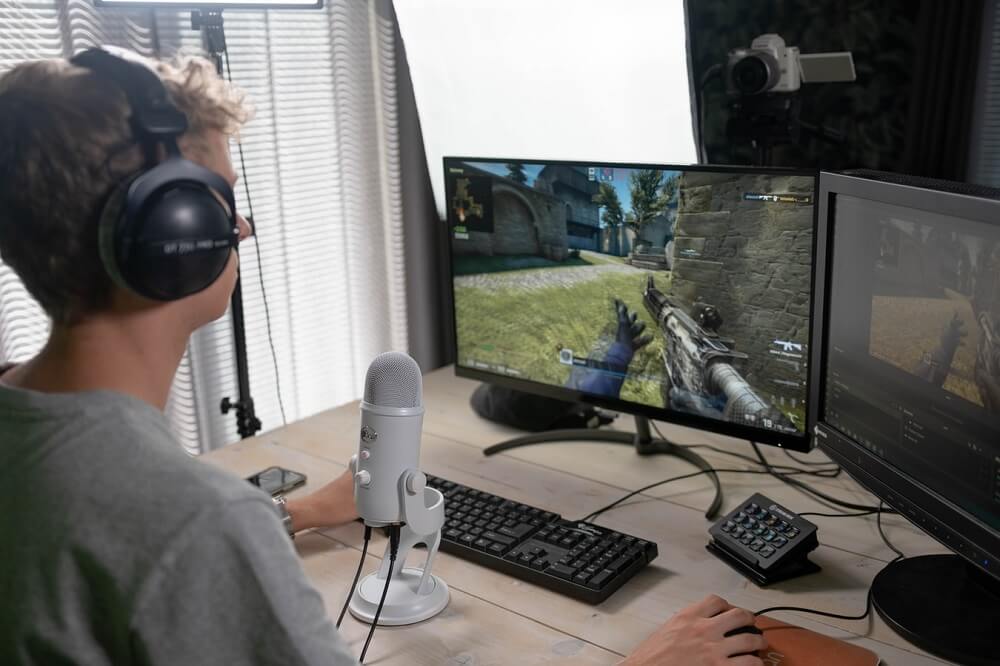 Image of a man streaming an FPS game with two monitors, a microphone and a camera in front of him