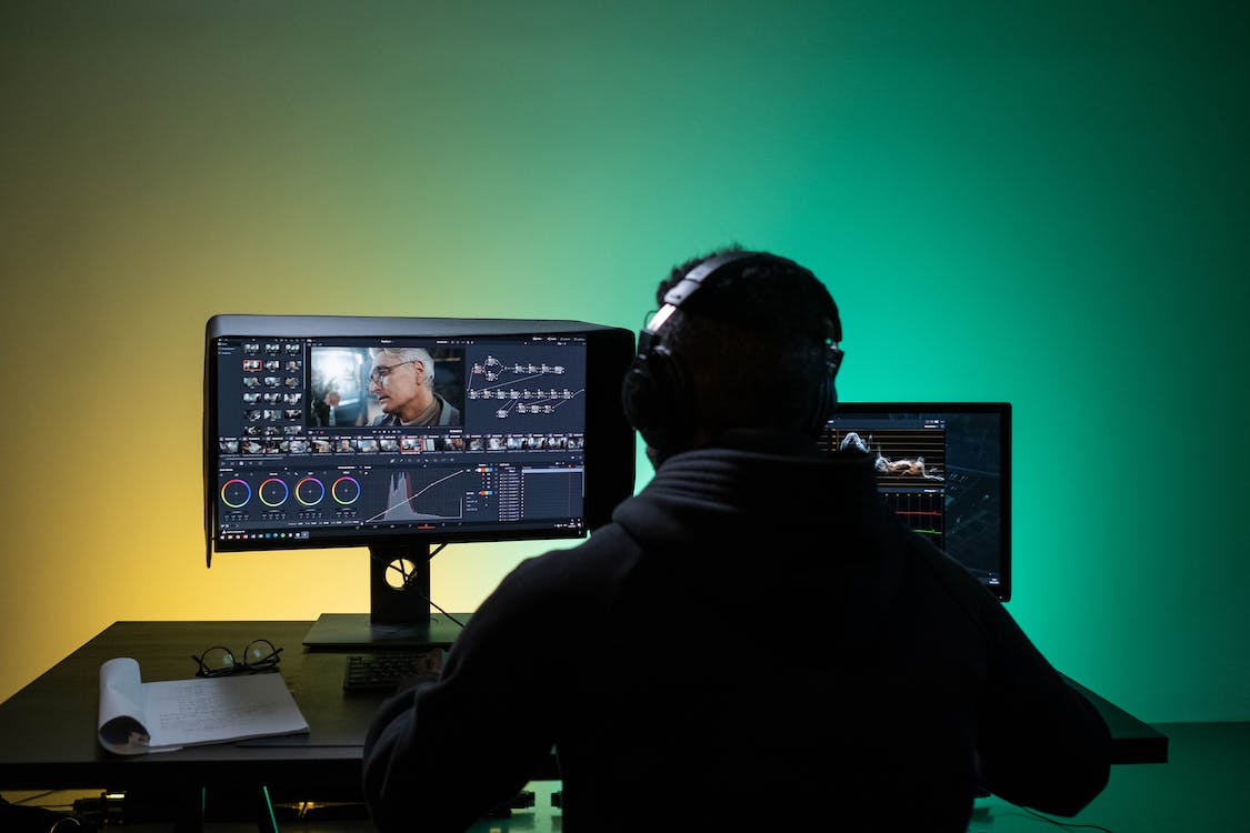 Photo of a man sitting at a desk with two monitors in front of him editing and rendering a video