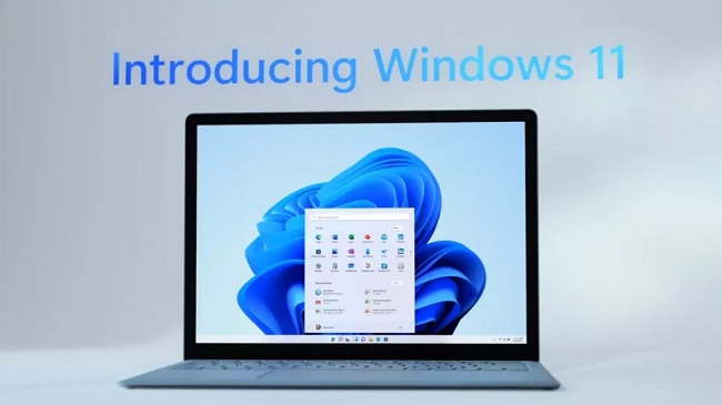 Promotional image for Microsoft Windows 11 showing a laptop open with Windows 11 on screen with the words 'Introducing Windows 11' on the wall behind it
